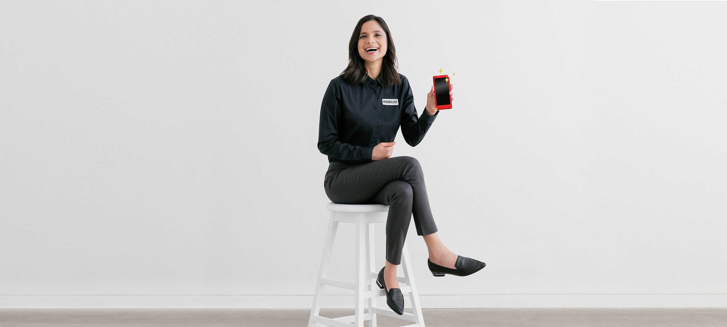 A laughing Mobilist woman showing a phone perching on a white chair